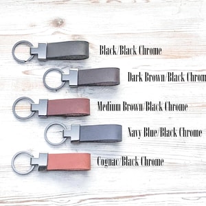 Leather keychains sitting on a white washed wood table. Black, dark brown, medium brown, navy blue and cognac leather keychains with black chrome hardware,