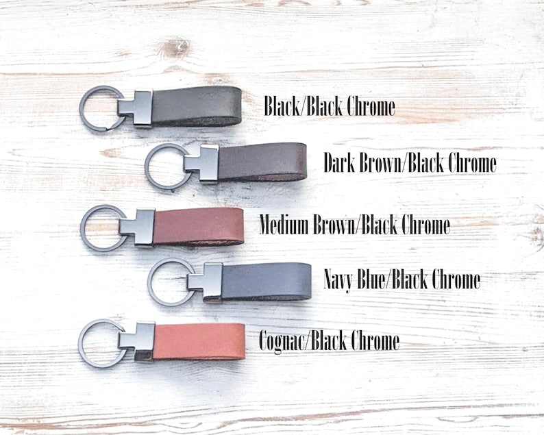 Leather keychains sitting on a white washed wood table. Black, dark brown, medium brown, navy blue and cognac leather keychains with black chrome hardware,