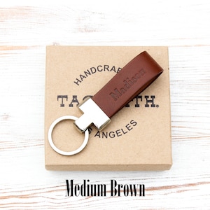 May include: A medium brown leather keyring featuring a silver-toned metal hardware attachment and a circular split ring. The leather strap is embossed with the name Madison. The keyring rests on a square cardboard box with printed text.