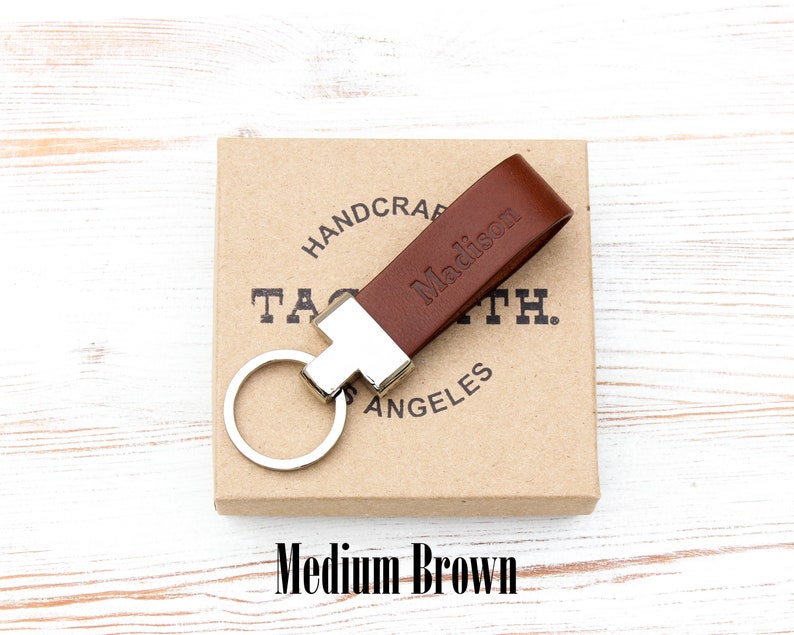 May include: A medium brown leather keyring featuring a silver-toned metal hardware attachment and a circular split ring. The leather strap is embossed with the name Madison. The keyring rests on a square cardboard box with printed text.