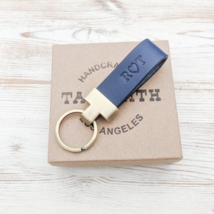 May include: A navy blue leather keyring featuring a gold-toned metal clasp and a circular ring. The leather strap is embossed with the letters R and T separated by a heart symbol. The keyring is positioned on a square cardboard box.