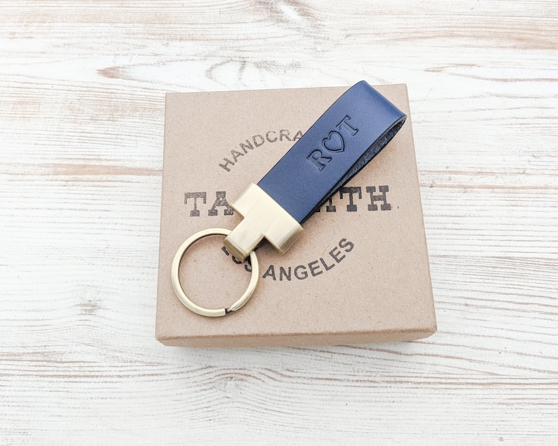 May include: A navy blue leather keyring featuring a gold-toned metal clasp and a circular ring. The leather strap is embossed with the letters R and T separated by a heart symbol. The keyring is positioned on a square cardboard box.