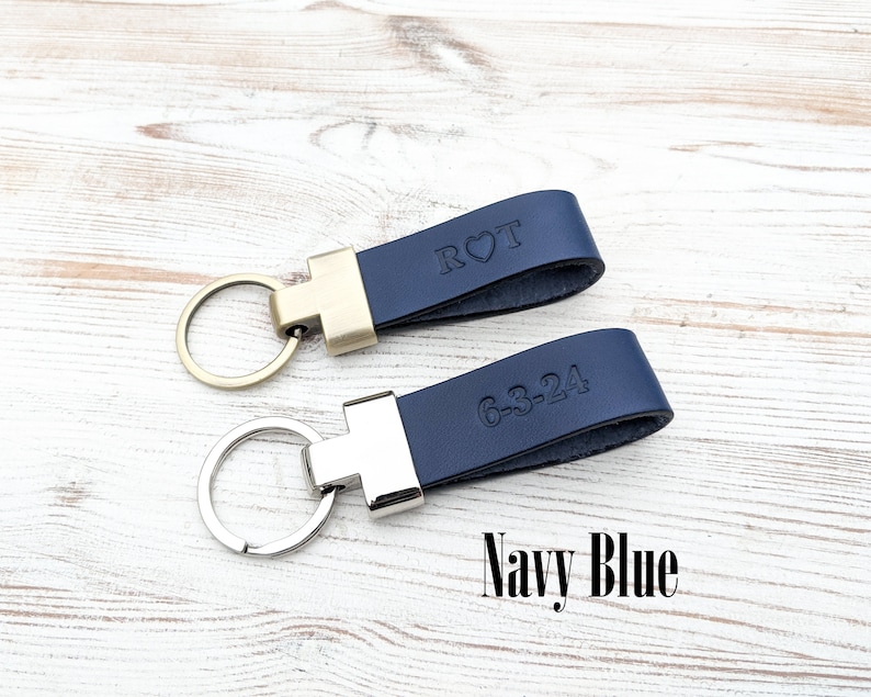 May include: Two navy blue leather keychains with metal hardware. One keychain features a gold-tone ring and the embossed text R heart T. The other keychain features a silver-tone ring and the embossed text 6-3-24. The text Navy Blue is visible.