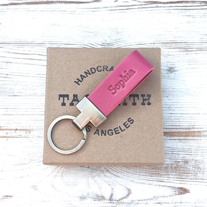 May include: A magenta leather keyring featuring a metallic silver-toned hardware attachment and a circular split ring. The leather strap is embossed with the name Sophia in a clear, sans-serif font, providing a personalised touch to the accessory.