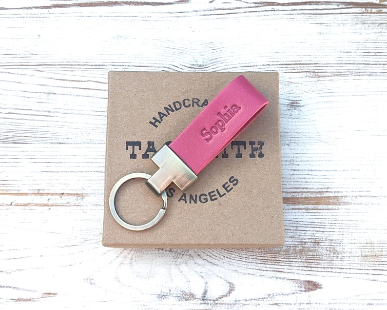 May include: A magenta leather keyring featuring a metallic silver-toned hardware attachment and a circular split ring. The leather strap is embossed with the name Sophia in a clear, sans-serif font, providing a personalised touch to the accessory.