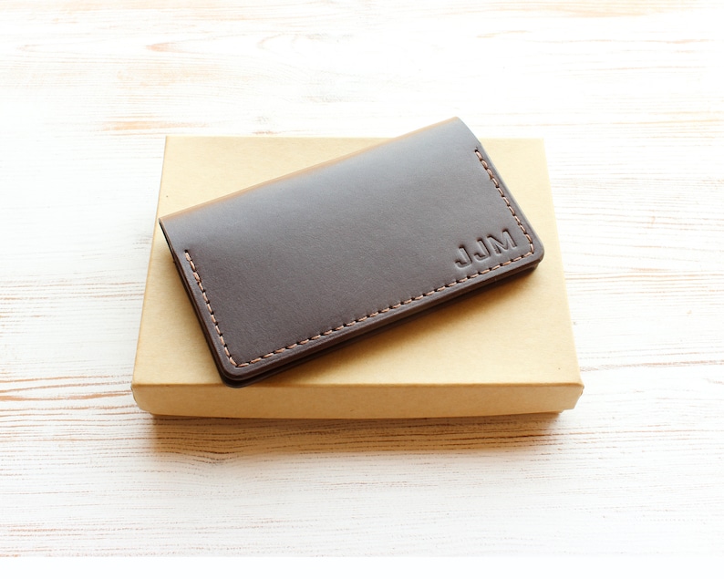 A dark brown leather business card holder sits on top of a kraft gift box, which is placed on a white washed wooden table. The card case is personalized with three debossed initials.