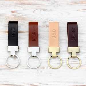 May include: Four leather keychains in black, medium brown, light tan, and dark brown. Each features a metal attachment and a circular ring. The leather straps are embossed with the names or initials DTM, Madison, J&D, and Hunter respectively.