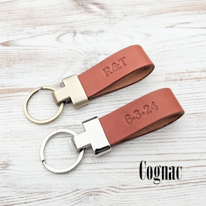 May include: Two cognac-coloured leather keychains with metal loops. One keychain features the embossed text R&T, while the other displays the date 6-3-24. The top keychain has a brass-toned metal connector, and the bottom one has a silver-toned connector.