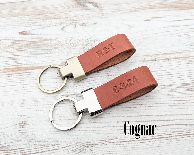 May include: Two cognac-coloured leather keychains with metal loops. One keychain features the embossed text R&T, while the other displays the date 6-3-24. The top keychain has a brass-toned metal connector, and the bottom one has a silver-toned connector.