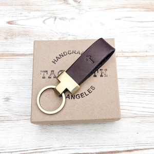 Dark brown leather keychain with antique brass hardware sits on top of a Tagsmith gift box, which sits on top of whitewashed wood grain. This custom keychain is personalized with a religious cross symbol debossed into the leather with a hot stamping machine.