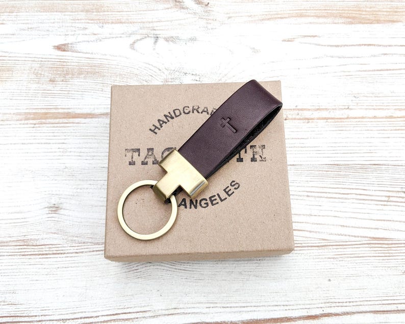Dark brown leather keychain with antique brass hardware sits on top of a Tagsmith gift box, which sits on top of whitewashed wood grain. This custom keychain is personalized with a religious cross symbol debossed into the leather with a hot stamping machine.