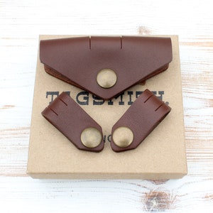 May include: A brown leather wallet and two matching leather accessories with brass snap closures. The items are displayed on a cardboard box with the text "TAGSMITH" printed on it. The leather has a smooth texture.