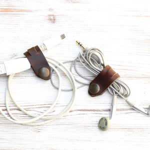 May include: Two brown leather cable organizers with brass snap closures. One organizer holds a white USB cable, and the other holds a gray headphone cable. The organizers are on a white wooden surface.