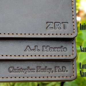 Three dark brown leather business card holders are stacked, each with different customization. One card case is personalized with initials, one with a name, and one with a long name.