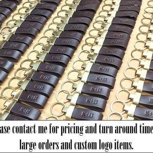 May include: A large collection of dark brown leather keychains arranged in rows. Each keychain features a brass-toned metal ring and a rectangular leather strap embossed with the letters K+H. The text below reads: Please contact me for pricing and turn around time on large orders and custom logo items.