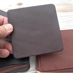 May include: A square, dark brown leather coaster is held by a hand, showcasing its smooth texture and rounded corners. Other coasters in various shades of brown and black are stacked in the background, suggesting a set of drinkware accessories.