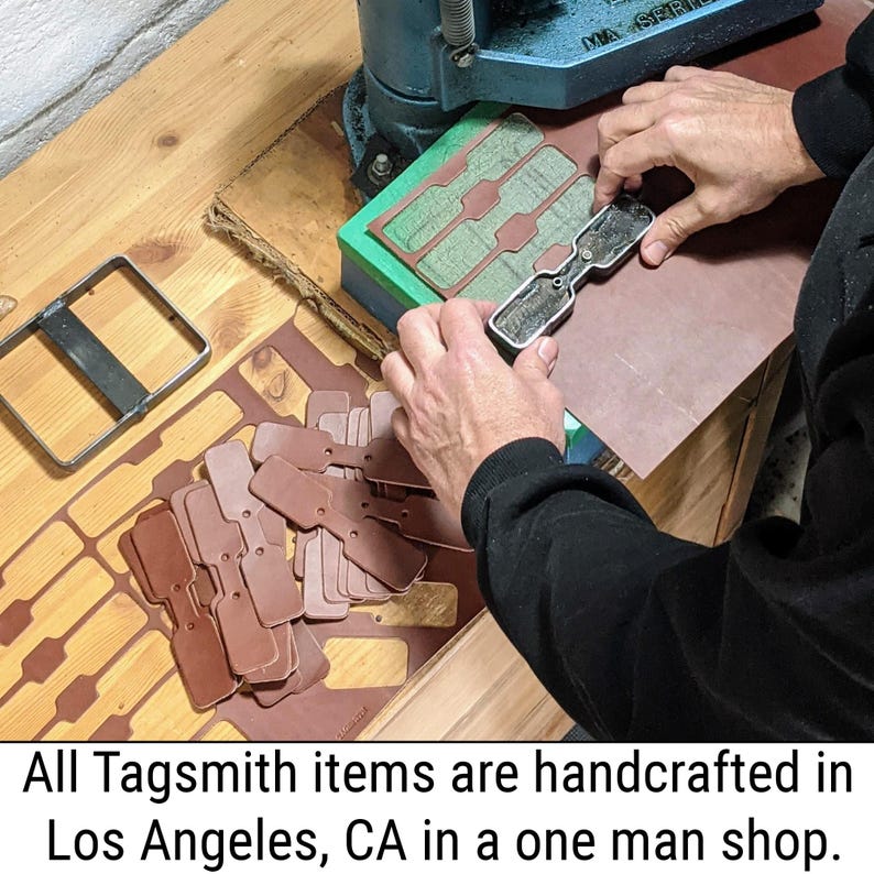 May include: A metal die press tool is used to cut brown leather tags from a sheet. The process involves a silver-coloured metal die, a blue press machine, and a pile of finished rectangular leather tags with rounded edges and a centre hole.