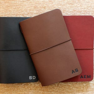May include: Three leather journals in black, brown, and red, each with a personalized initial. The journals are closed with an elastic band. The initials "SD", "AS", and "AEM" are visible on the covers.