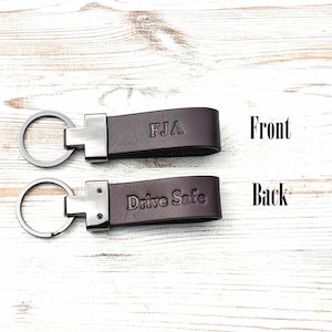May include: Two dark brown leather keychains with silver-toned metal hardware and circular key rings. The top keychain is embossed with the letters FJA on the front, while the bottom keychain features the embossed text Drive Safe on the back.