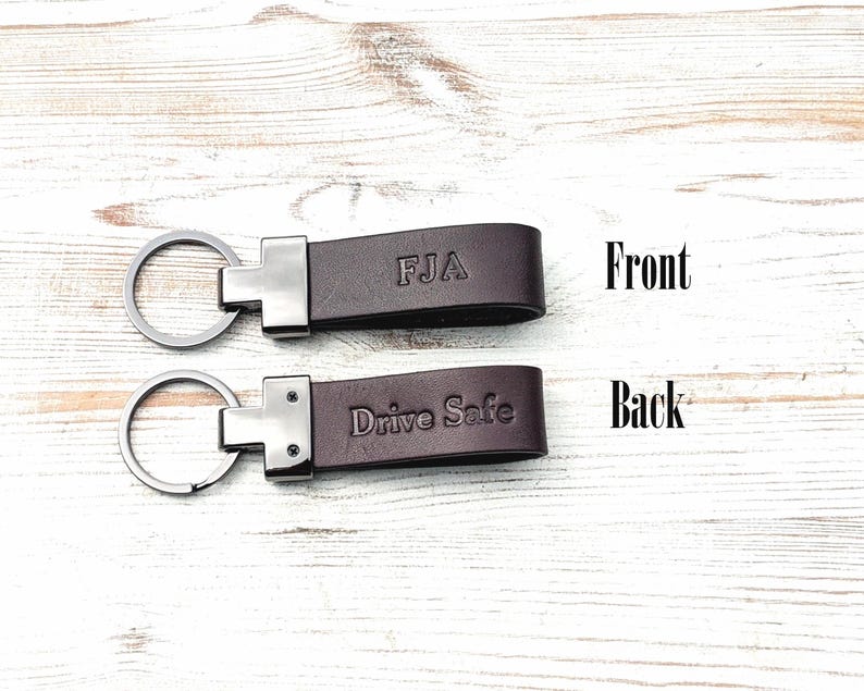 May include: Two dark brown leather keychains with silver-toned metal hardware and circular key rings. The top keychain is embossed with the letters FJA on the front, while the bottom keychain features the embossed text Drive Safe on the back.