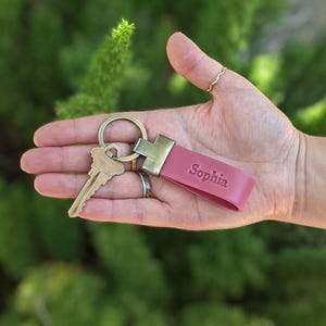 May include: A pink leather keyring featuring a brass-toned metal clasp and a split ring holding a single brass-coloured key. The leather strap is embossed with the name Sophia in a clear, sans-serif font, providing a simple and functional accessory.