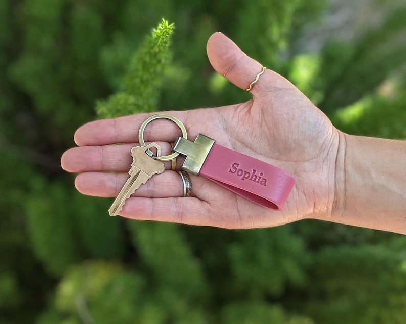 May include: A pink leather keyring featuring a brass-toned metal clasp and a split ring holding a single brass-coloured key. The leather strap is embossed with the name Sophia in a clear, sans-serif font, providing a simple and functional accessory.