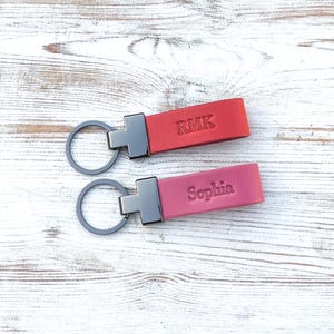 May include: Two rectangular leather keychains with dark metal hardware and circular rings. The top keychain is red and embossed with the letters RMK. The bottom keychain is a dusty rose colour and embossed with the name Sophia in a serif font.
