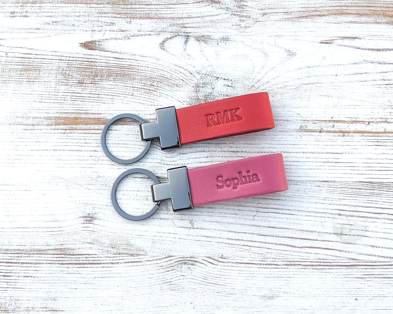 May include: Two rectangular leather keychains with dark metal hardware and circular rings. The top keychain is red and embossed with the letters RMK. The bottom keychain is a dusty rose colour and embossed with the name Sophia in a serif font.