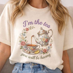 May include: Cream-colored t-shirt featuring a floral tea party design. The graphic includes a teapot, teacup, and the text "I'm the tea you will be having" in purple and green. The design is surrounded by a floral wreath.