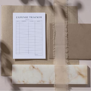 May include: A white printable expense tracker sheet with the words "Expense Tracker" at the top. The sheet is on a brown corrugated cardboard surface with a white marble surface below it.