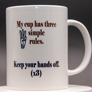 May include: White ceramic coffee mug with a curved handle. The mug features the text "My cup has three simple rules. Keep your hands off. (x3)" in brown and black lettering. A hand gesture graphic is also included.