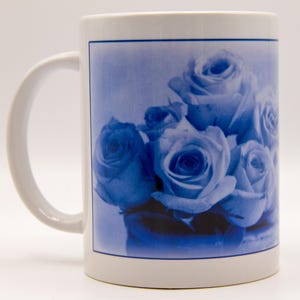 May include: White ceramic mug featuring a blue floral design of roses. The roses are clustered together, with a soft, watercolor-like effect. The mug has a white handle and a blue border.