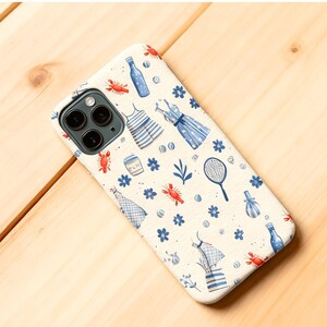 Może przedstawiać: Kremowe etui na telefon z akwarelowym wzorem ilustracji o tematyce letniej. Projekt zawiera niebieskie sukienki, homary, rakiety tenisowe i butelki. Etui jest przeznaczone do smartfona z trzema obiektywami.