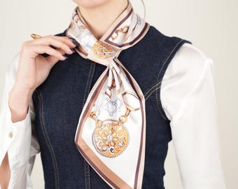 May include: A cream-colored silk scarf with a detailed equestrian-themed design. The scarf features gold, brown, and blue accents, with a repeating pattern of horse bits, chains, and medallions. The scarf is tied around the neck, creating a stylish accessory.