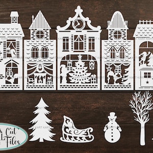 Op de afbeelding: Witte papieren uitsparingen van een kerstdorp met huizen, bomen, een sneeuwpop en een slee. De huizen hebben gedetailleerde ramen, deuren en schoorstenen. De tekst "DIGITAL Cut Files" is inbegrepen.