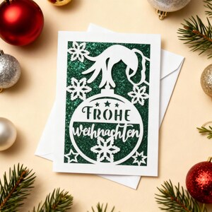 Peut inclure: Une carte de No&euml;l blanche avec un fond vert paillet&eacute; et un motif d'ornement d&eacute;coup&eacute;. L'ornement porte le texte allemand "Frohe Weihnachten" et est entour&eacute; de flocons de neige et d'&eacute;toiles. La carte est pos&eacute;e sur une surface beige clair avec des d&eacute;corations de No&euml;l et des branches de pin.