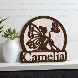 Può includere: Un'insegna in legno tagliata al laser con una silhouette di fata con ali di farfalla, che tiene in mano una farfalla. L'insegna è marrone con sfondo in legno chiaro e il nome "Camelia" in basso. Articolo decorativo.