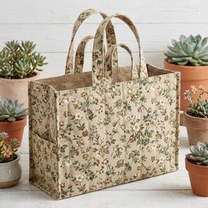 Puede incluir: Un bolso tote acolchado rectangular con base de tela beige y un estampado floral en tonos marrón, verde y blanco. El bolso incluye dos pares de asas de tela, una parte superior abierta estructurada y un bolsillo lateral en el exterior.