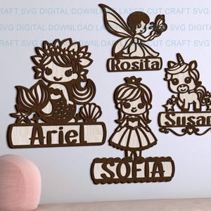 May include: Wooden cutouts of a mermaid, fairy, princess, and unicorn, each with a name. The mermaid is named Ariel, the fairy Rosita, the princess Sofia, and the unicorn Susan. The cutouts are brown and have a light wood background.