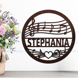 Pode incluir: Decoração de parede de madeira castanha escura em forma circular, com notas musicais e o nome "STEPHANIA". Uma forma de coração na parte inferior. Um cesto de flores e uma estatueta no fundo.