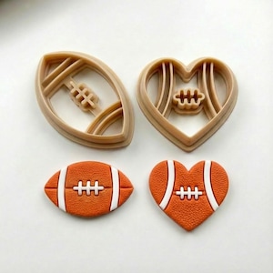 May include: Set of four football-themed cookie cutters and cookies. Two tan plastic cutters are shaped like a football and a heart. Two orange cookies are shaped like a football and a heart, with white stripes and stitching.