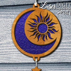May include: Wooden celestial ornament featuring a sun and crescent moon design. The sun is in the center of the ornament, with the moon surrounding it. The moon is filled with blue glitter. The ornament is hanging by a white string.