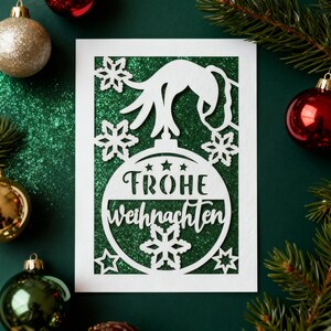 Peut inclure: Carte de No&euml;l blanche avec un motif d&eacute;coup&eacute; repr&eacute;sentant une boule de No&euml;l avec l'inscription "Frohe Weihnachten" et des flocons de neige. La carte est pos&eacute;e sur un fond vert paillet&eacute;, avec des d&eacute;corations de No&euml;l et des branches de pin.
