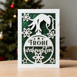以下が含まれることがあります： 「FROHE WEIHNACHTEN」の文字と雪の結晶が描かれたクリスマスオーナメントを特徴とする、切り絵デザインの白いクリスマスカード。背景は緑色のグリッター。カードは長方形で、テクスチャ加工された表面です。
