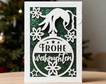May include: A white Christmas card with a cut-out design featuring a Christmas ornament with the words "FROHE WEIHNACHTEN" and snowflakes. The background is green glitter. The card is rectangular and has a textured surface.