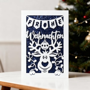 以下が含まれることがあります： 白いクリスマスカード。トナカイとドイツ語の「Frohe Weihnachten」（メリークリスマス）のフレーズがカットアウトデザインで描かれています。背景は濃い青色のグリッター。お祝いの挨拶。