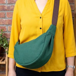 Può includere: Una borsa a tracolla a forma di mezzaluna di colore verde scuro realizzata in tessuto a tinta unita. La borsa presenta un'ampia tracolla intrecciata marrone che si collega agli angoli superiori del corpo curvo. Design semplice e arrotondato.