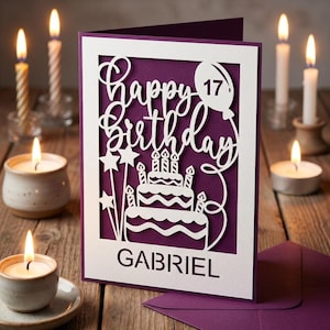 Op de afbeelding: Een witte lasergesneden verjaardagskaart met de woorden "Happy Birthday" en "Gabriel" op een paarse achtergrond. De kaart toont een taart, sterren en een ballon met het nummer 17. Brandende kaarsen omringen de kaart op een houten oppervlak.