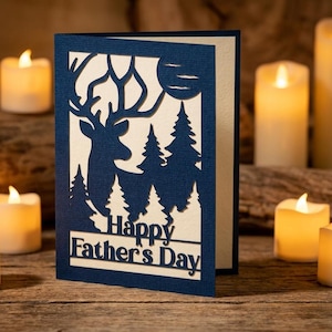 May include: A navy blue laser-cut greeting card featuring a silhouette of a deer, pine trees, and a moon against a cream-colored background. The text "Happy Father's Day" is cut out at the bottom of the card, which stands upright.