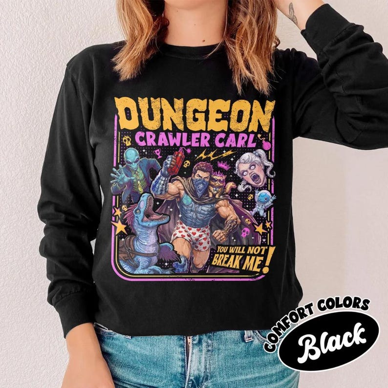 May include: A black long-sleeve shirt featuring a graphic print with the text "DUNGEON CRAWLER CARL" and "YOU WILL NOT BREAK ME!". The illustration depicts a central muscular figure in heart-patterned boxers surrounded by various fantasy characters.
